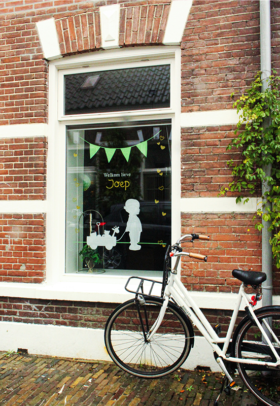 Huis met geparkeerde witte fiets voor. Raamtekening van vlaggetjes boven een jongen met knuffels in een kar. 